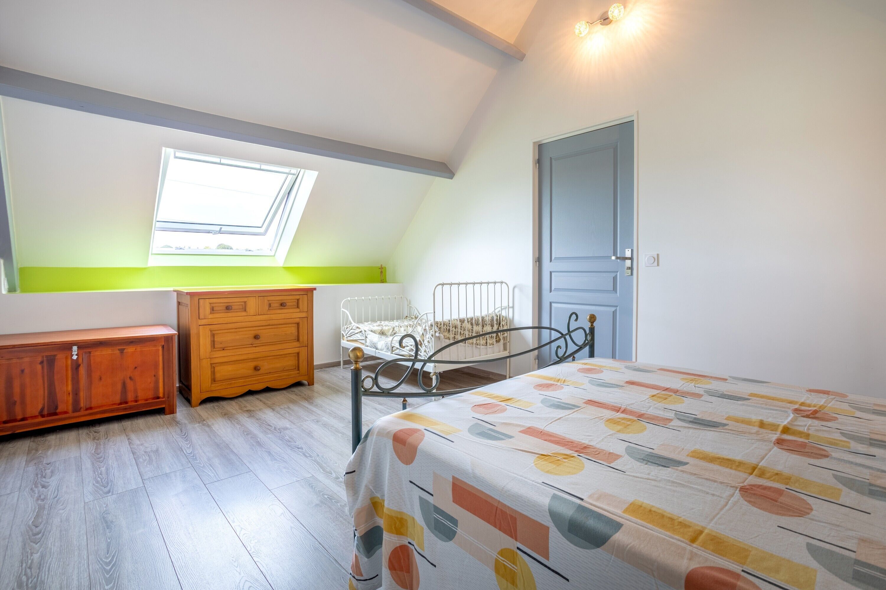 A bedroom with a sloped ceiling, a skylight, a bed with a patterned bedspread, a wooden dresser, and a metal bed frame.