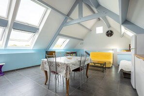 Un comedor con una mesa y sillas, un sofá amarillo, un reloj en la pared y una zona de cocina con un refrigerador.