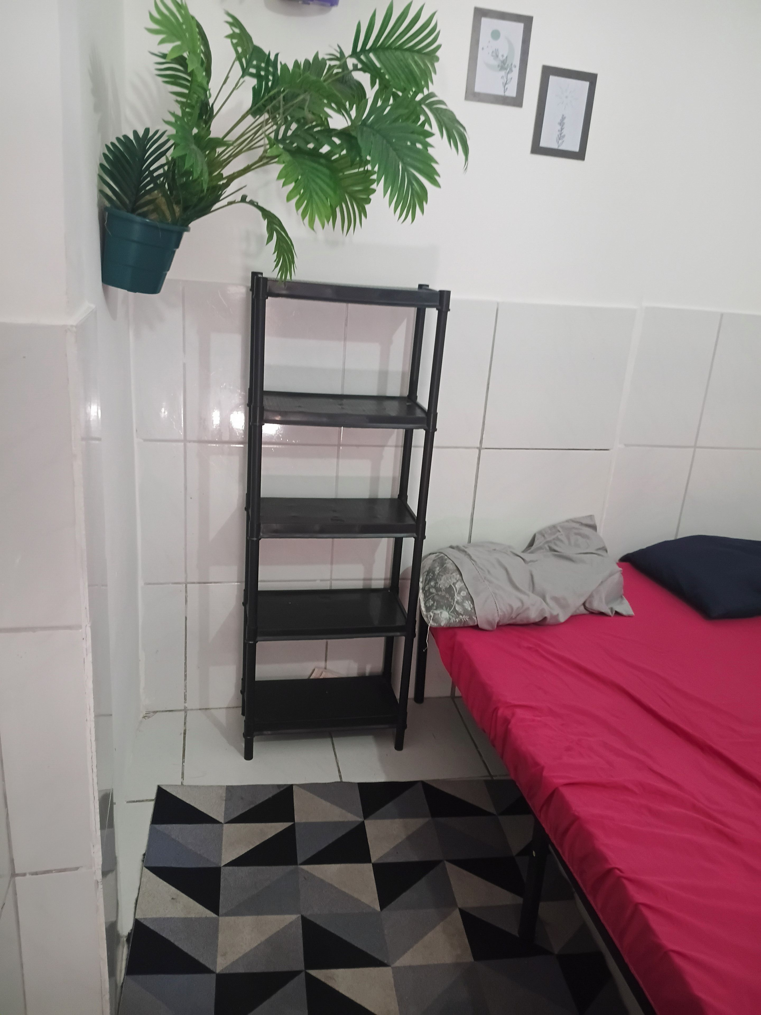Una habitación con cama, una estantería metálica negra, un tapete estampado, una planta en maceta y una pared con cuadros.