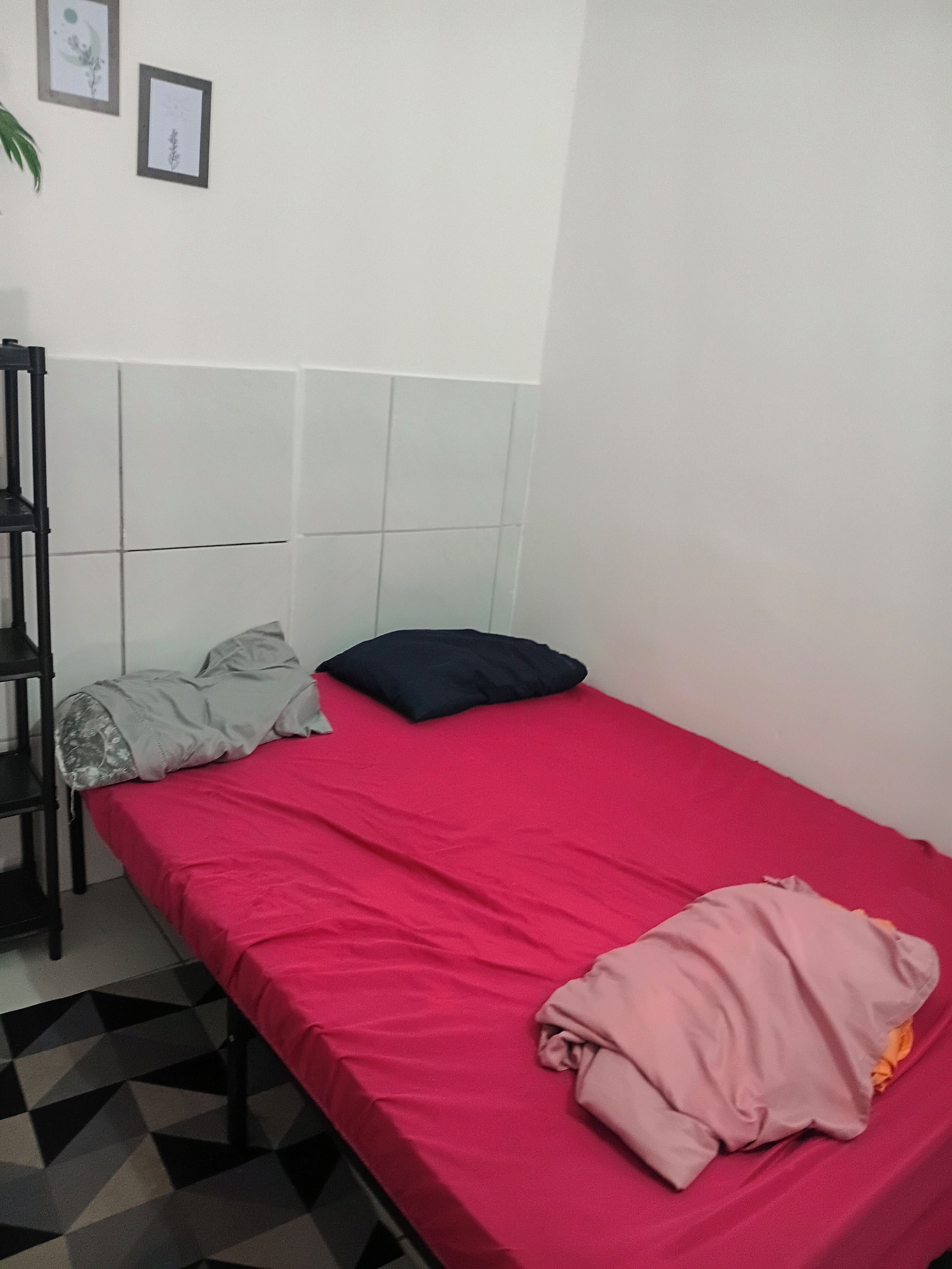 Una habitación pequeña con cama individual, colcha roja, estante negro y piso estampado.