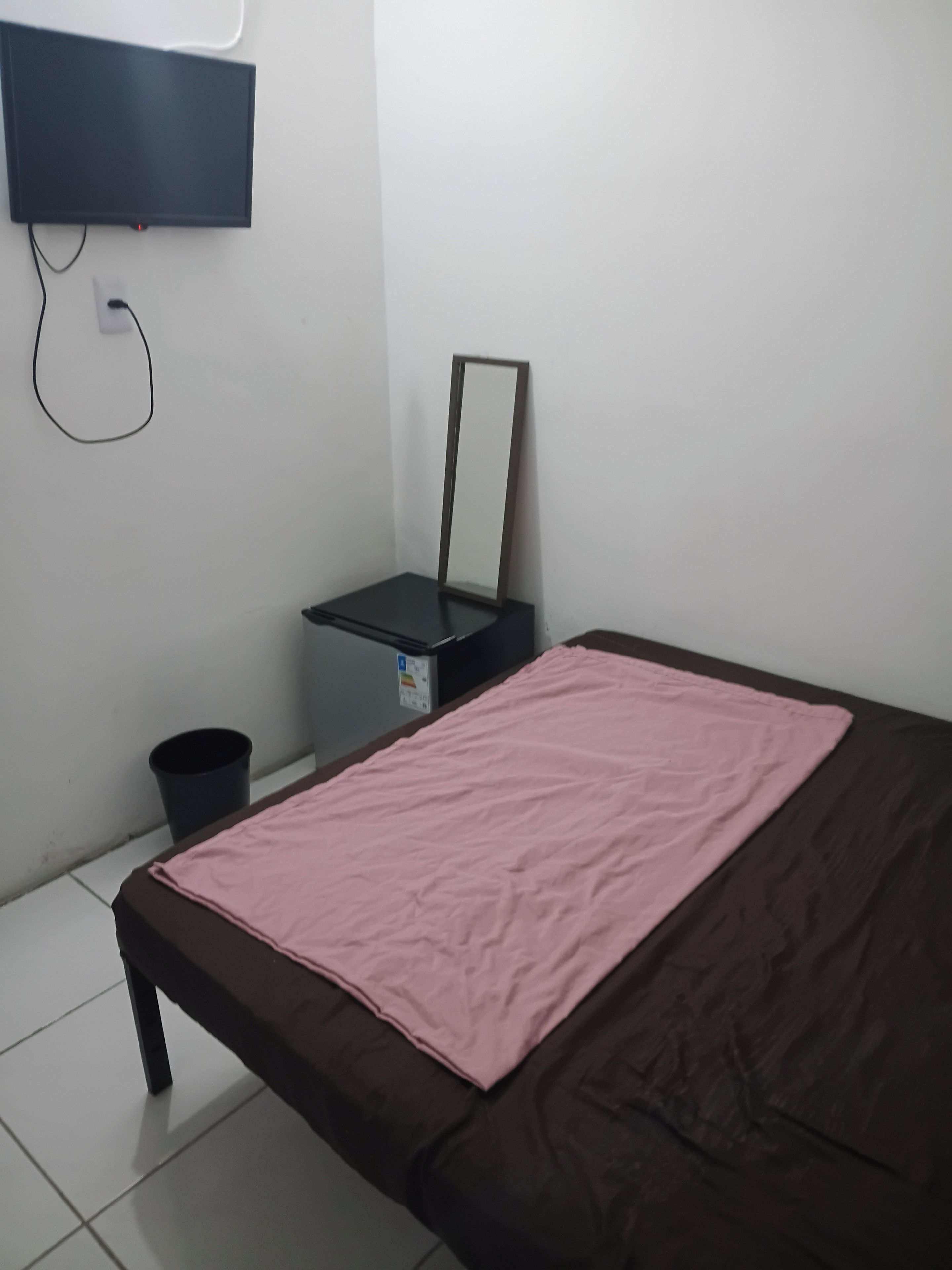 Una habitación con una cama, una mesita, una silla, un cubo de basura y un televisor en la pared.