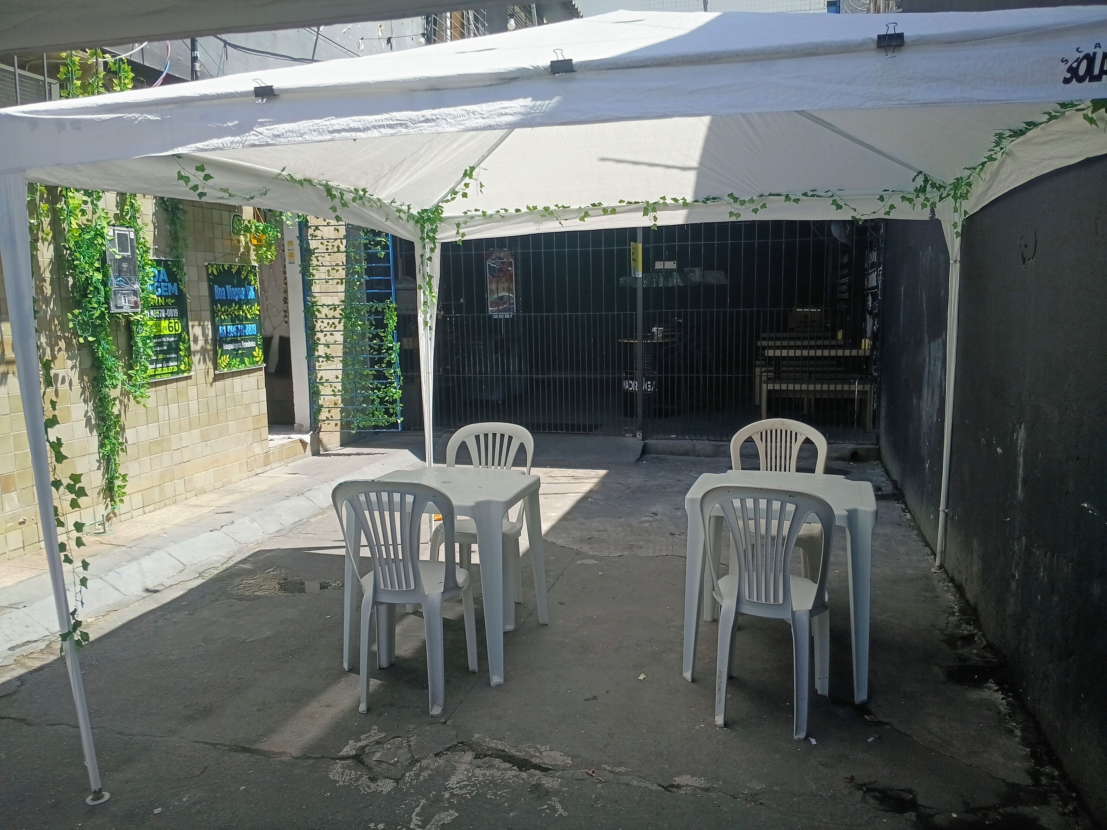 Un área de estar al aire libre con techo, con sillas y mesas de plástico, rodeada de muros con vegetación y una entrada con reja.