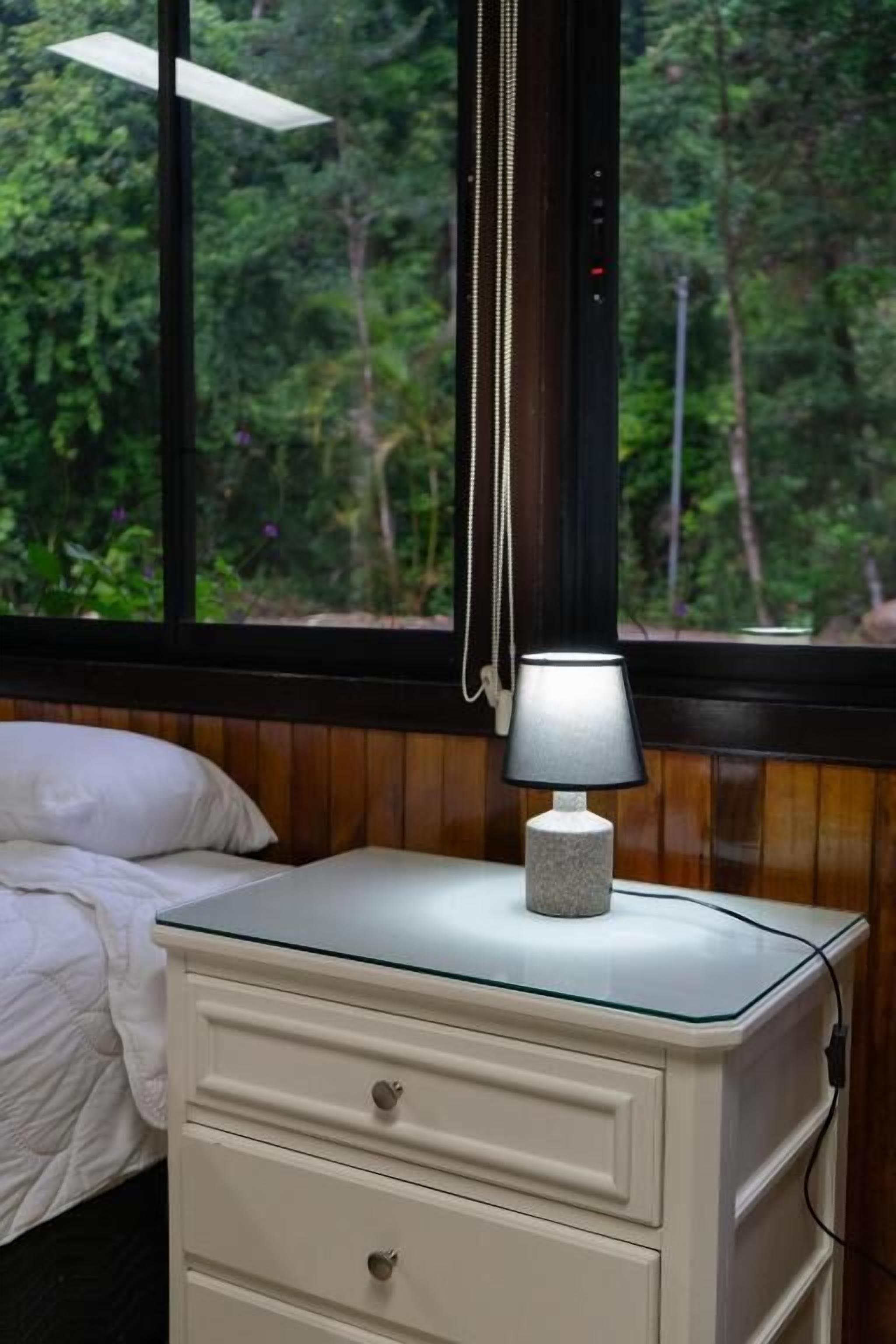 Un lit avec une literie blanche, une lampe de chevet, une tête de lit en bois et une fenêtre donnant sur un paysage verdoyant.