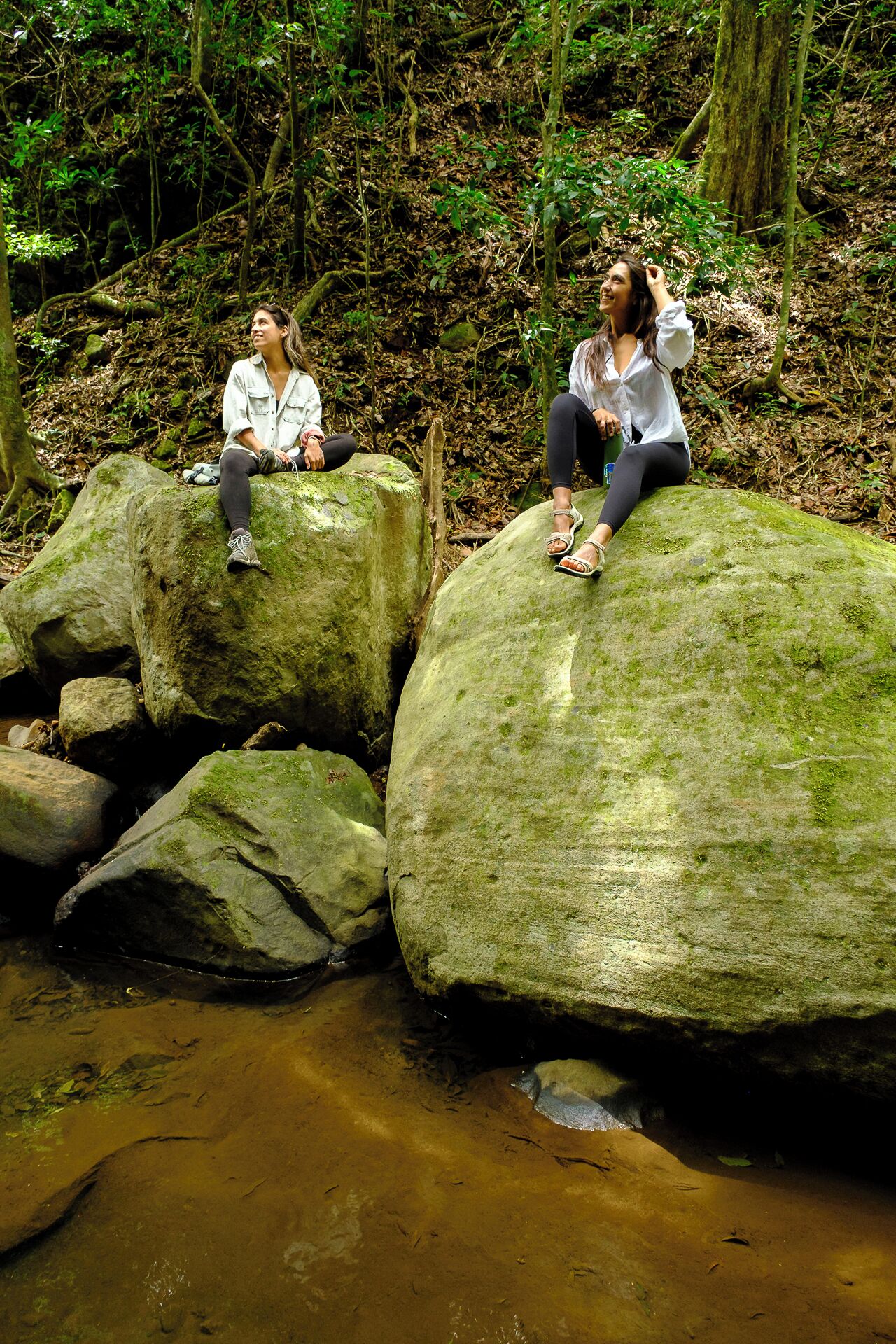 Two individuals sitting on moss-covered rocks by a stream in a forested area.