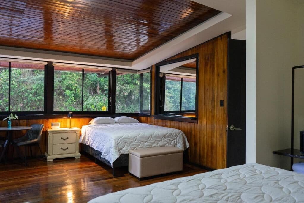 Un dormitorio con techo de madera, un ventanal con vistas a árboles, una cama con colcha blanca, una mesita de noche y una silla.