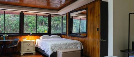 Un dormitorio con techo de madera, un ventanal con vistas a árboles, una cama con colcha blanca, una mesita de noche y una silla.
