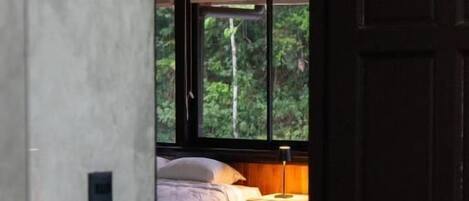 A bedroom with a bed, bedside table, and a window with a view of greenery.