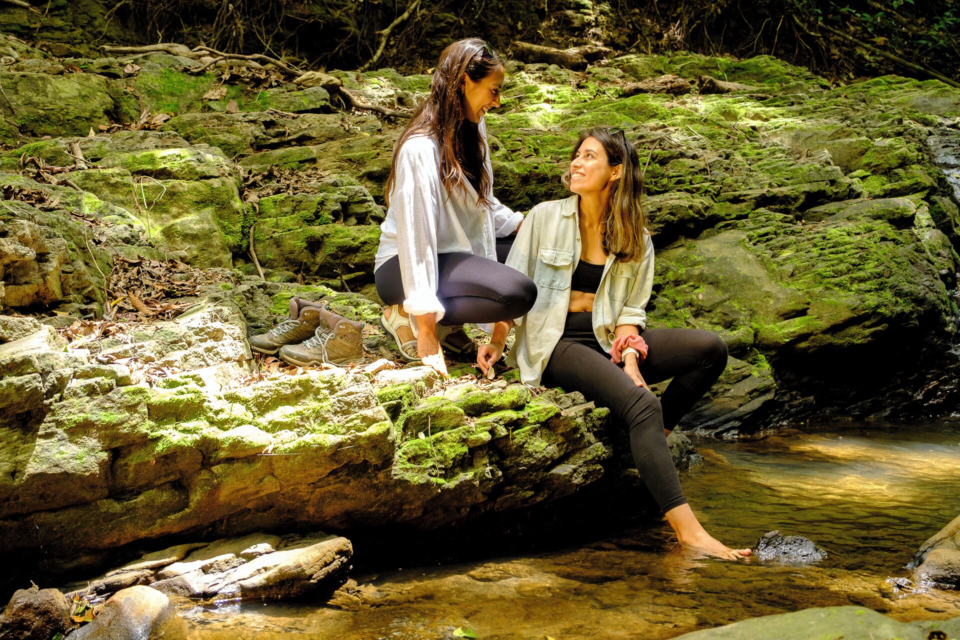 Two people sitting on a moss-covered rock by a clear stream.