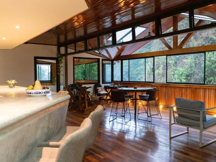 Um quarto espaçoso com teto de madeira, janelas amplas, área de jantar com cadeiras e mesa, e bancada de cozinha com frutas.