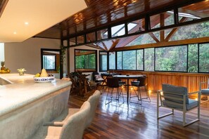 Una habitación amplia con techo de madera, ventanas grandes, una zona de comedor con sillas y mesa, y una encimera de cocina con fruta.