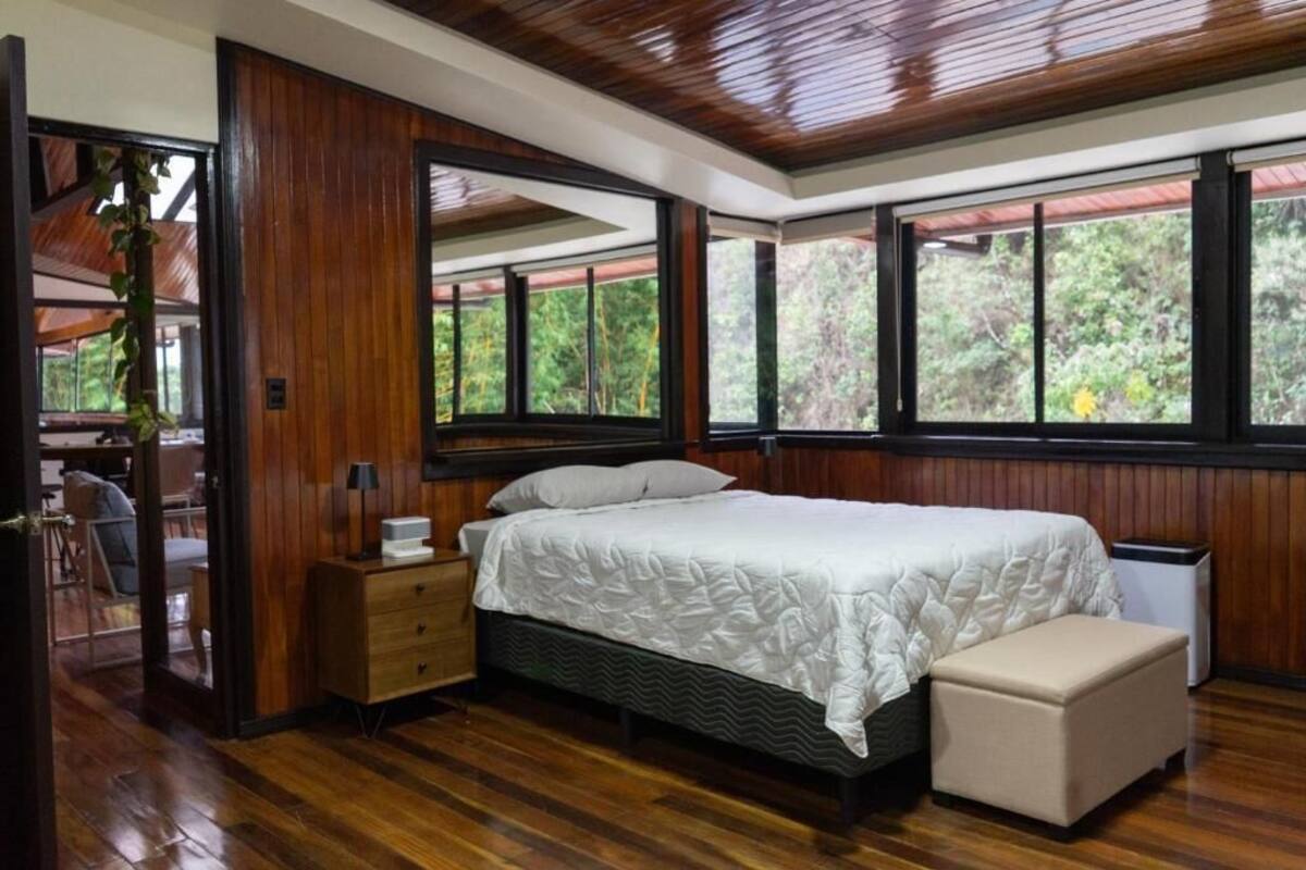 A bedroom with a bed, bedside table, bench, and large windows with a view of greenery.