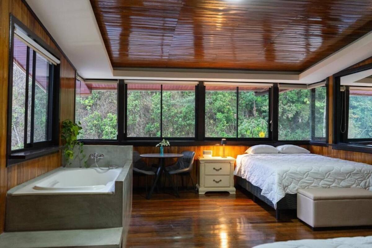 A bedroom with a large window, a bathtub, a bedside table, and a bed.