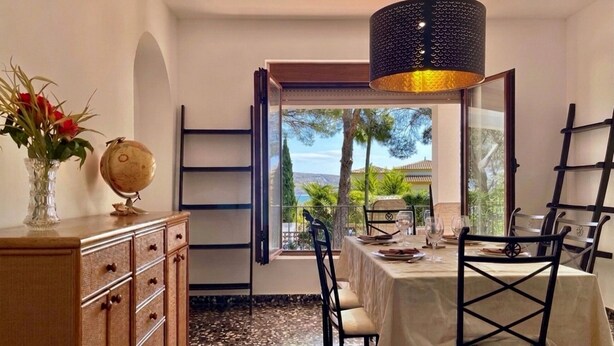 Comedor con mesa lista para dos, un globo en un mueble y vista a palmeras y un edificio a través de una ventana.