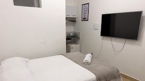 En snyggt bäddad säng med en hoprullad handduk, en tv som är monterad på väggen och ett litet skåp med toalettartiklar.