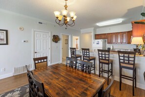 A dining area with a wooden table and chairs, a chandelier, and a kitchen in the background. - Ocean Villa 1802 | Oversized Balcony + Pool (Panama City Beach)