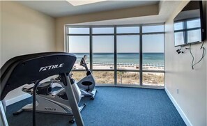 A room with a treadmill, elliptical, and large windows with a beach view. - Ocean Villa 1802 | Oversized Balcony + Pool (Panama City Beach)