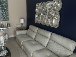 A beige leather sofa against a dark wall, a round side table with a lamp, and a decorative metal wall piece.