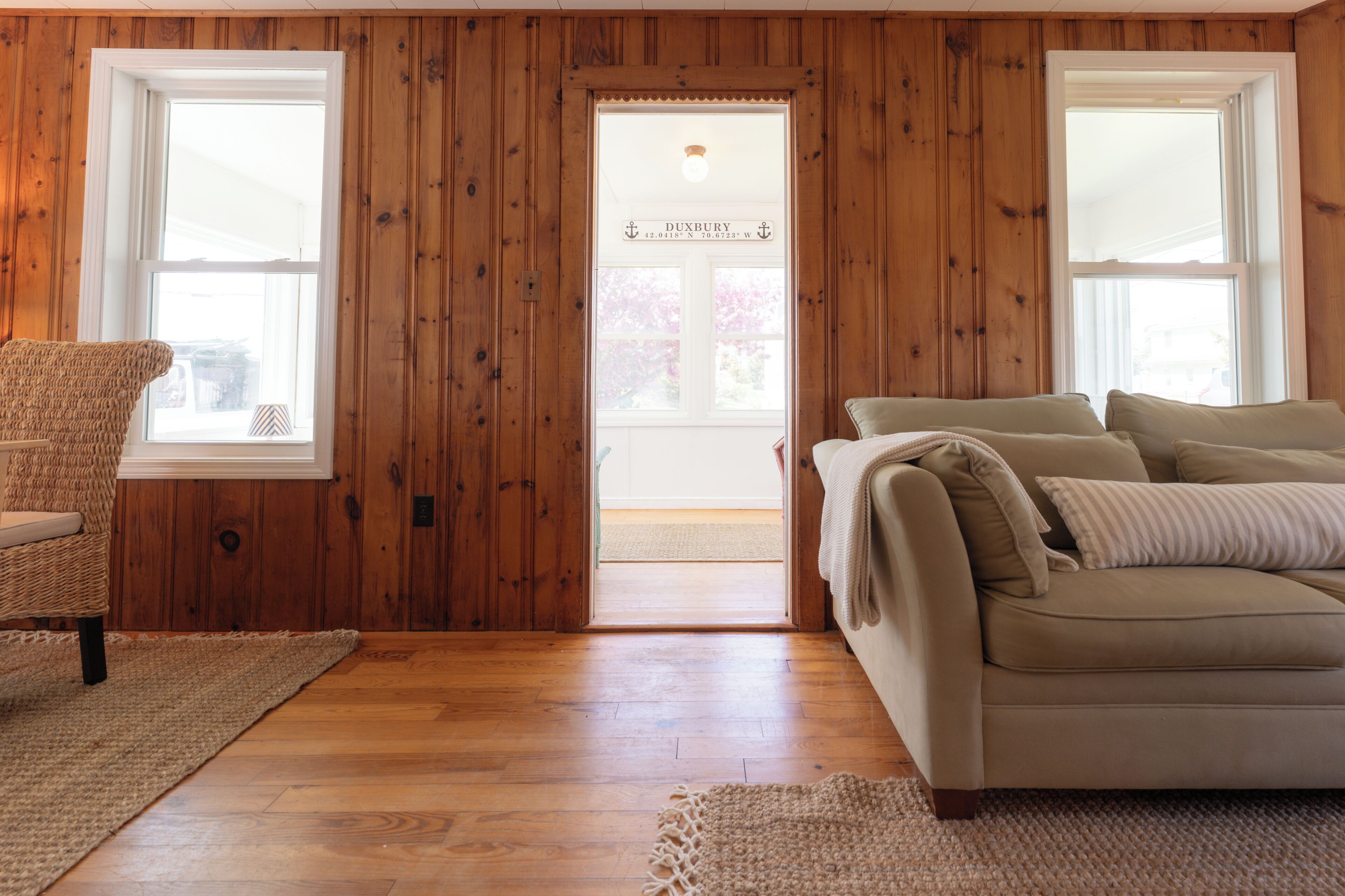 A room with wooden walls, a beige sofa, a wicker chair, and a door leading outside.