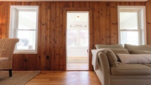 Una habitación con paredes de madera, un sofá color crema, una silla de mimbre y una puerta que da al exterior.