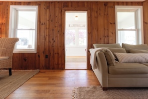 Ein Zimmer mit Holzwänden, einem beigen Sofa, einem Korbstuhl und einer Tür, die nach draußen führt.