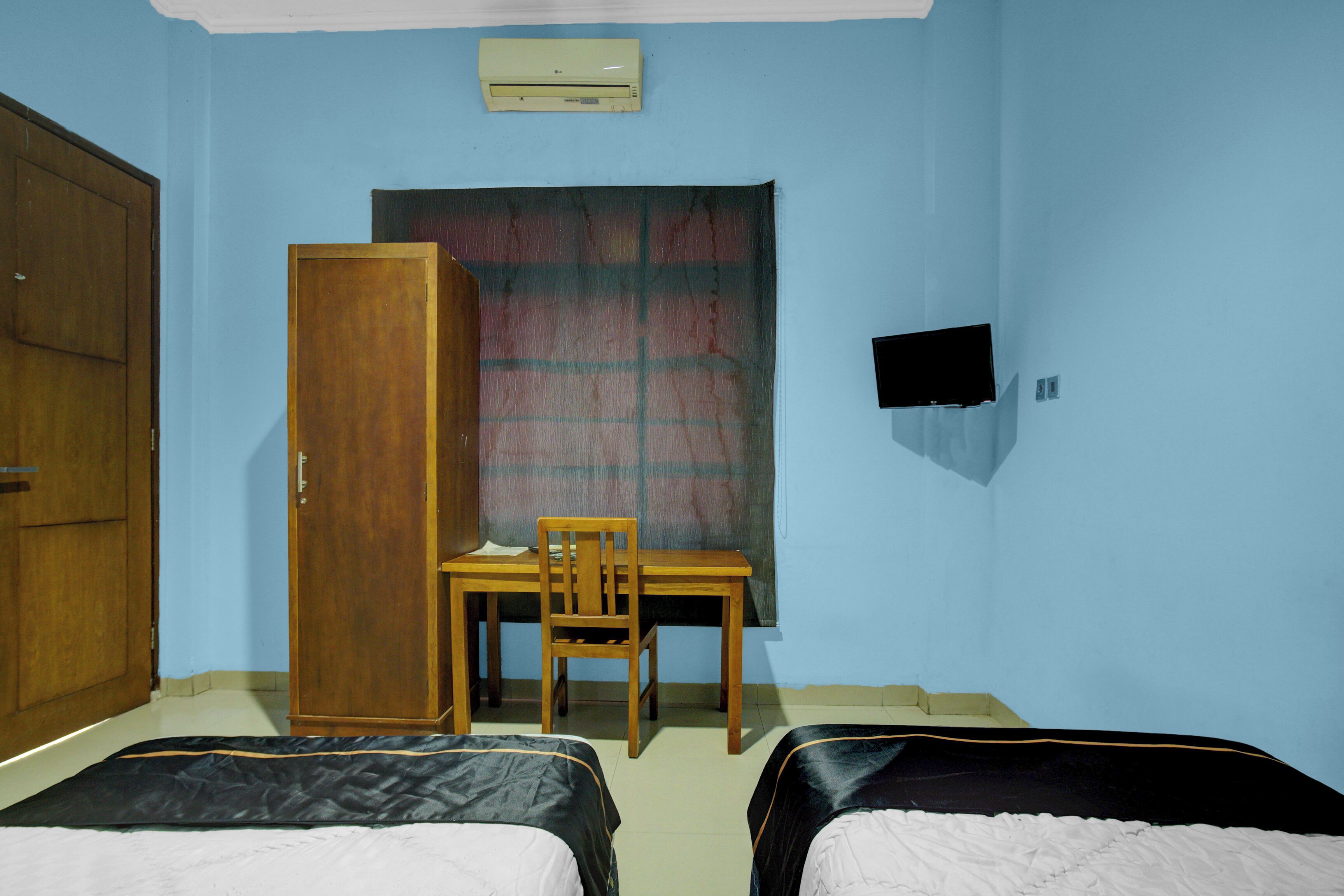 A room with two beds, a wooden wardrobe, a desk with a chair, and a television on the wall.