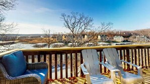 Una terraza con dos sillas Adirondack, una silla de mimbre y vistas a un pueblo con edificios y árboles.