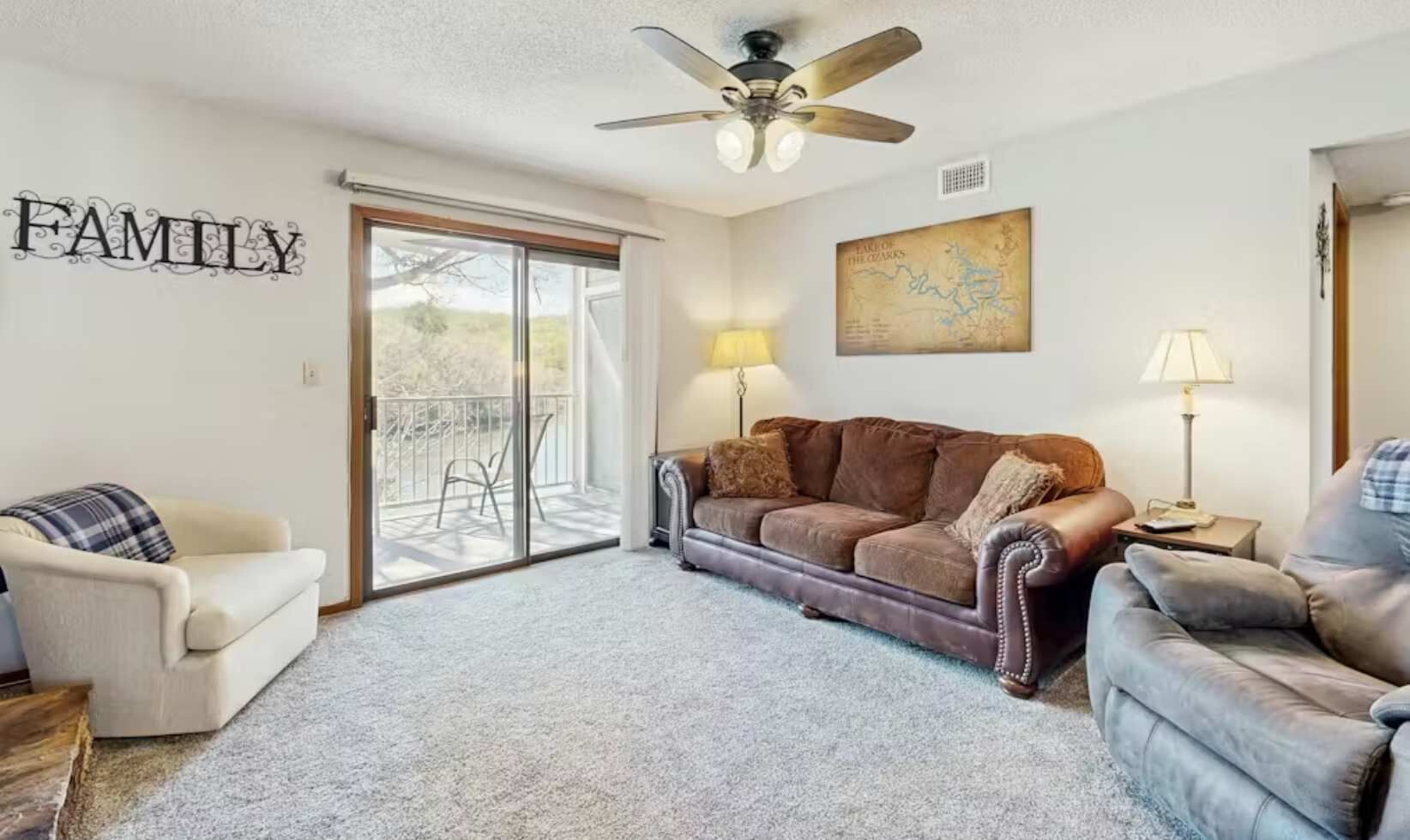 A living room with a brown sofa, a white sofa, a grey sofa, a ceiling fan, a map on the wall, and a sliding glass door leading to an outdoor area.