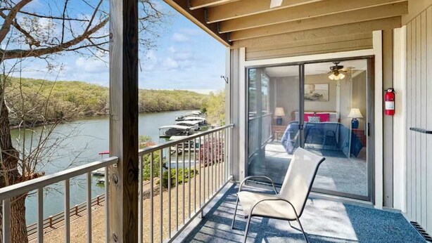 A balcony with a view of a river, a chair, and a room with a bed and a table. - Charming 2BR Condo - LAKE VIEW - Osage Beach (Osage Beach)
