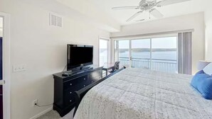 Une chambre à coucher avec un lit, un téléviseur posé sur une commode, un ventilateur de plafond et un balcon offrant une vue sur l’eau.