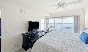 Une chambre à coucher avec un lit, un téléviseur posé sur une commode, un ventilateur de plafond et un balcon offrant une vue sur l’eau.