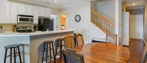 A kitchen with white cabinets, a microwave, a stove, and a refrigerator. There is a dining area with a wooden table and chairs.