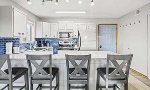 Una cocina moderna con armarios blancos, una isla central con taburetes y electrodomésticos como microondas, horno y refrigerador.