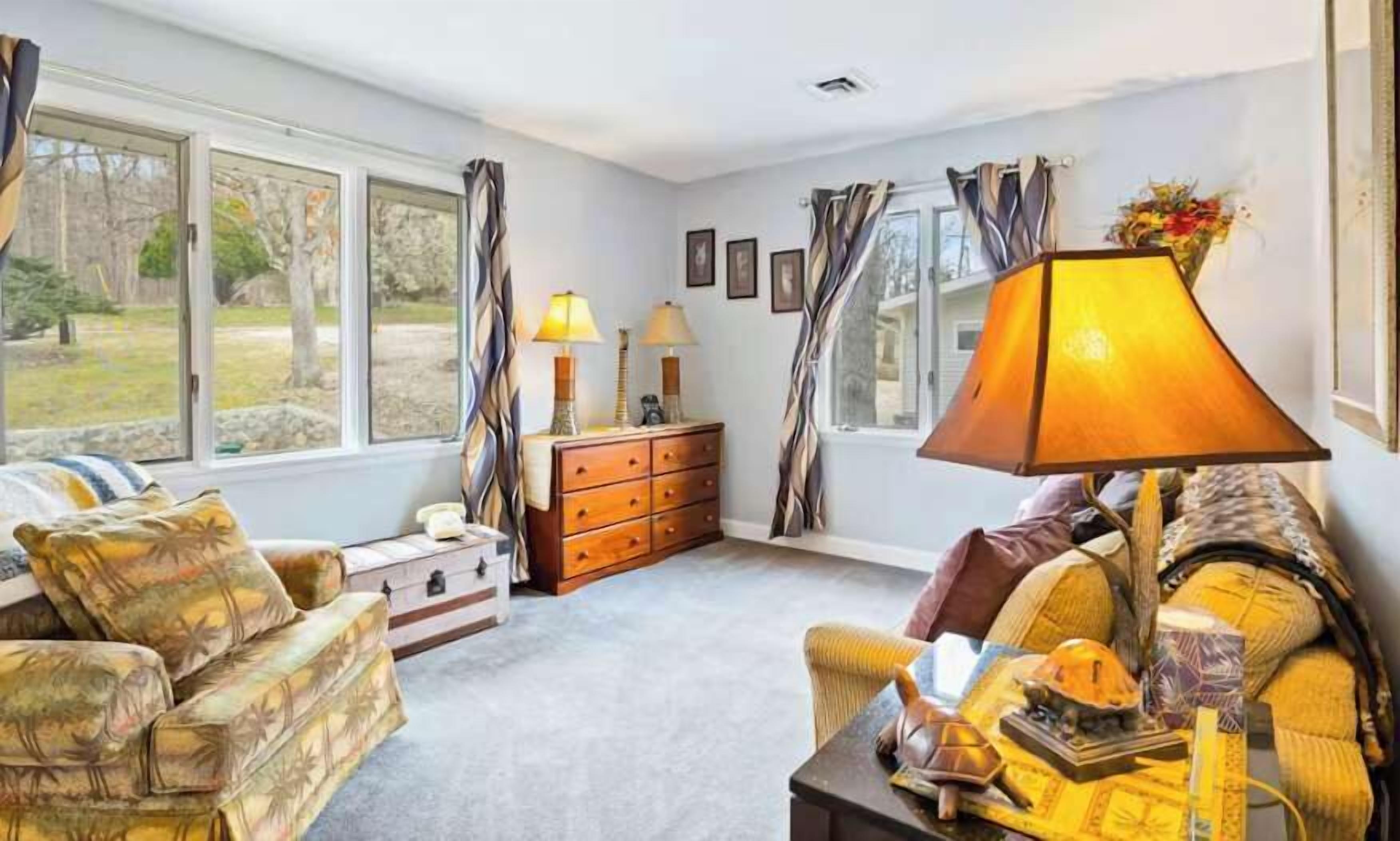 A living room with a sofa, a chair, a wooden dresser, and a lamp. There are windows with curtains, a fireplace, and a view of the outdoors.