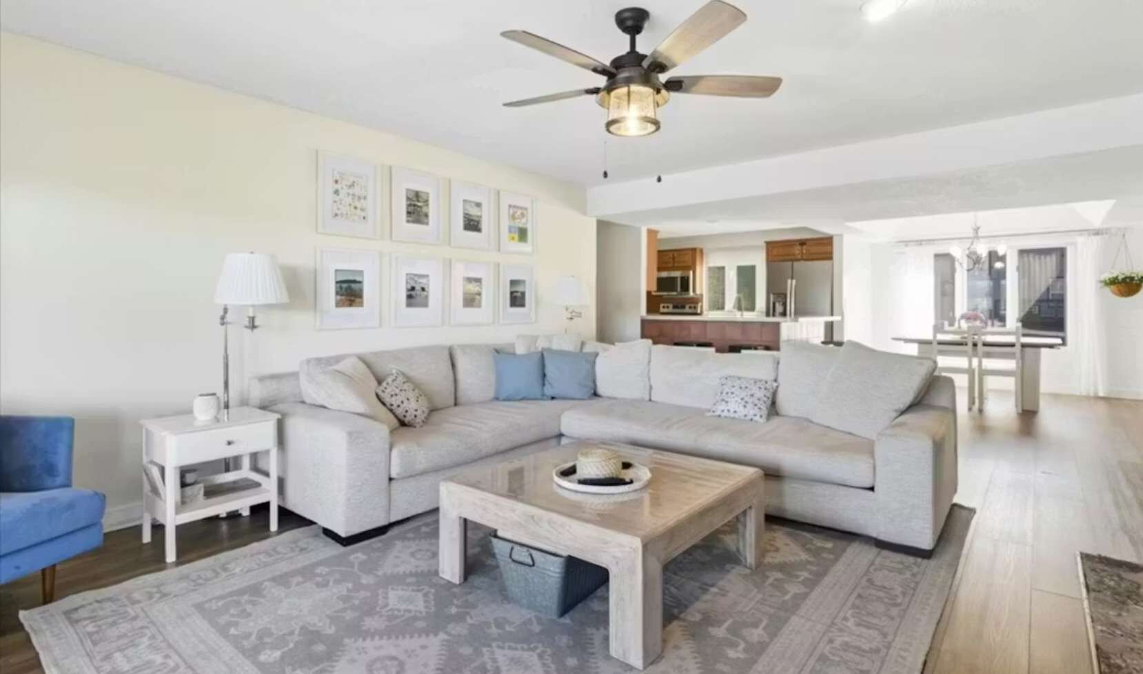Uma sala de estar com sofá seccional, mesa de centro, mesa lateral, abajur e quadros nas paredes.
