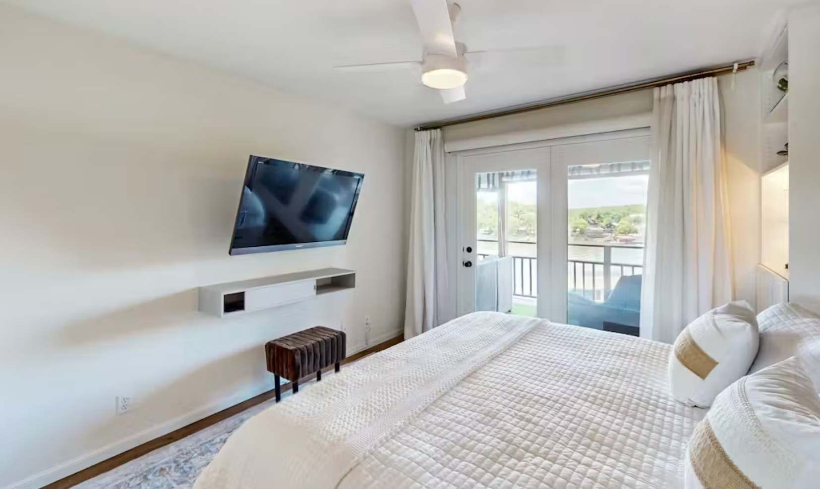 Une chambre d’hôtel avec un lit, un téléviseur fixé au mur, un banc et un balcon avec des rideaux.