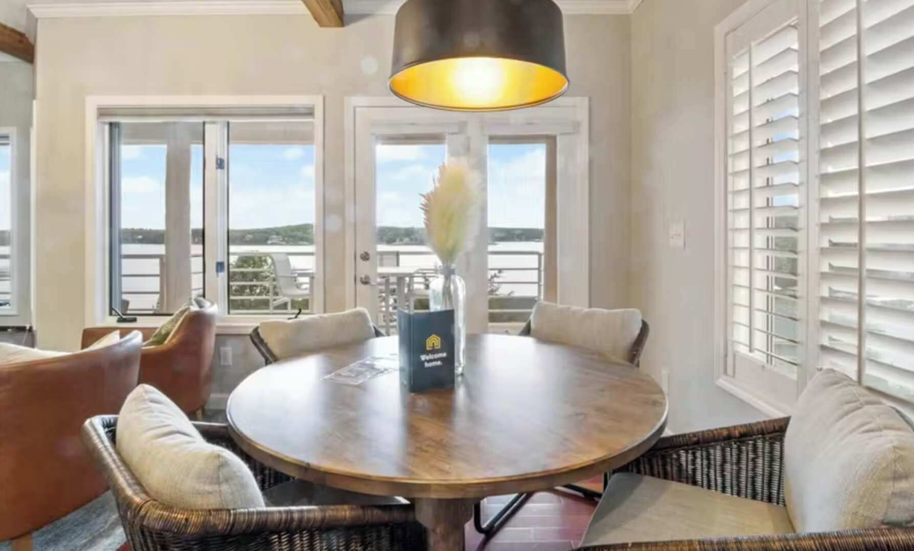 Un tavolo rotondo in legno con sedie, un vaso e un cartello di benvenuto in una stanza luminosa con ampie finestre e una vista.