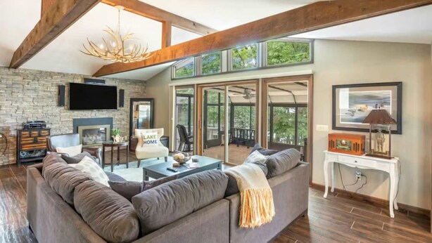 A spacious living room with a stone fireplace, a large sofa, and a television mounted on the wall. - Rustic 3BR Lake House - LAKE VIEW - Lake Ozark (Lake Ozark)