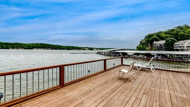 Eine Holzterrasse mit Geländer, die auf ein Gewässer blickt, zwei Liegestühle und ein Steg mit Gebäuden im Hintergrund.