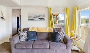 Un salon avec un canapé gris, des coussins à motifs, un tableau accroché au mur et une fenêtre ornée de rideaux jaunes.