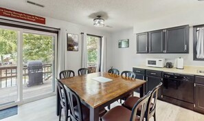 Un comedor con una mesa y sillas de madera, una cocina con armarios y electrodomésticos oscuros, y un vidrio corredero que da a una terraza exterior.