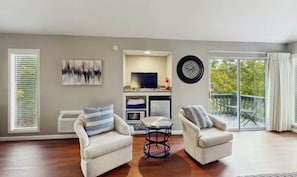 A living room with two armchairs, a small round table, a flat-screen TV, and a large window with blinds.