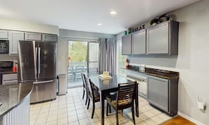 A modern kitchen with stainless steel appliances, a dining area with a table and chairs, and a view through a glass door to an outdoor area.