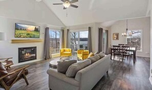 A spacious living room with a fireplace, a large sofa, yellow chairs, and a dining area with a table and chairs. - Serene 3BR House - LAKE VIEW - Margaritaville (Osage Beach)