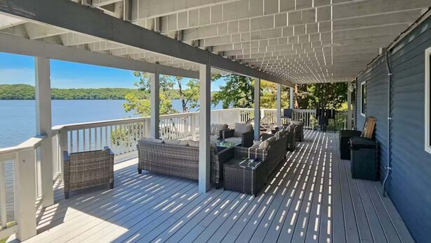 En inglasad veranda med utemöbler, utsikt över en sjö och en byggnad med en blå fasad.