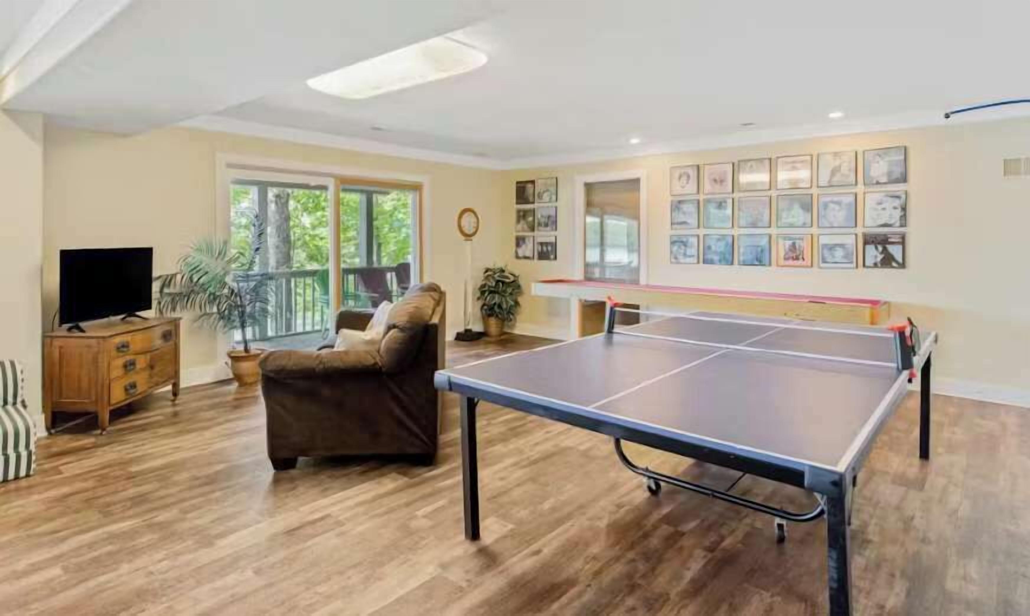 A living room with a ping pong table, a brown leather sofa, a TV on a wooden cabinet, and a large window with a view of greenery.
