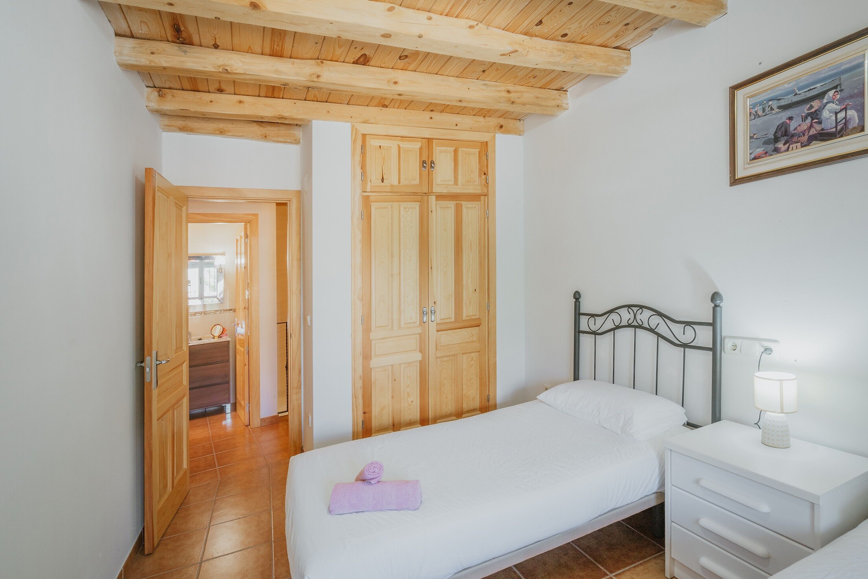 A bedroom with a wooden ceiling, a bed with a white mattress and a pink towel, a bedside table with a lamp, and a wooden wardrobe.