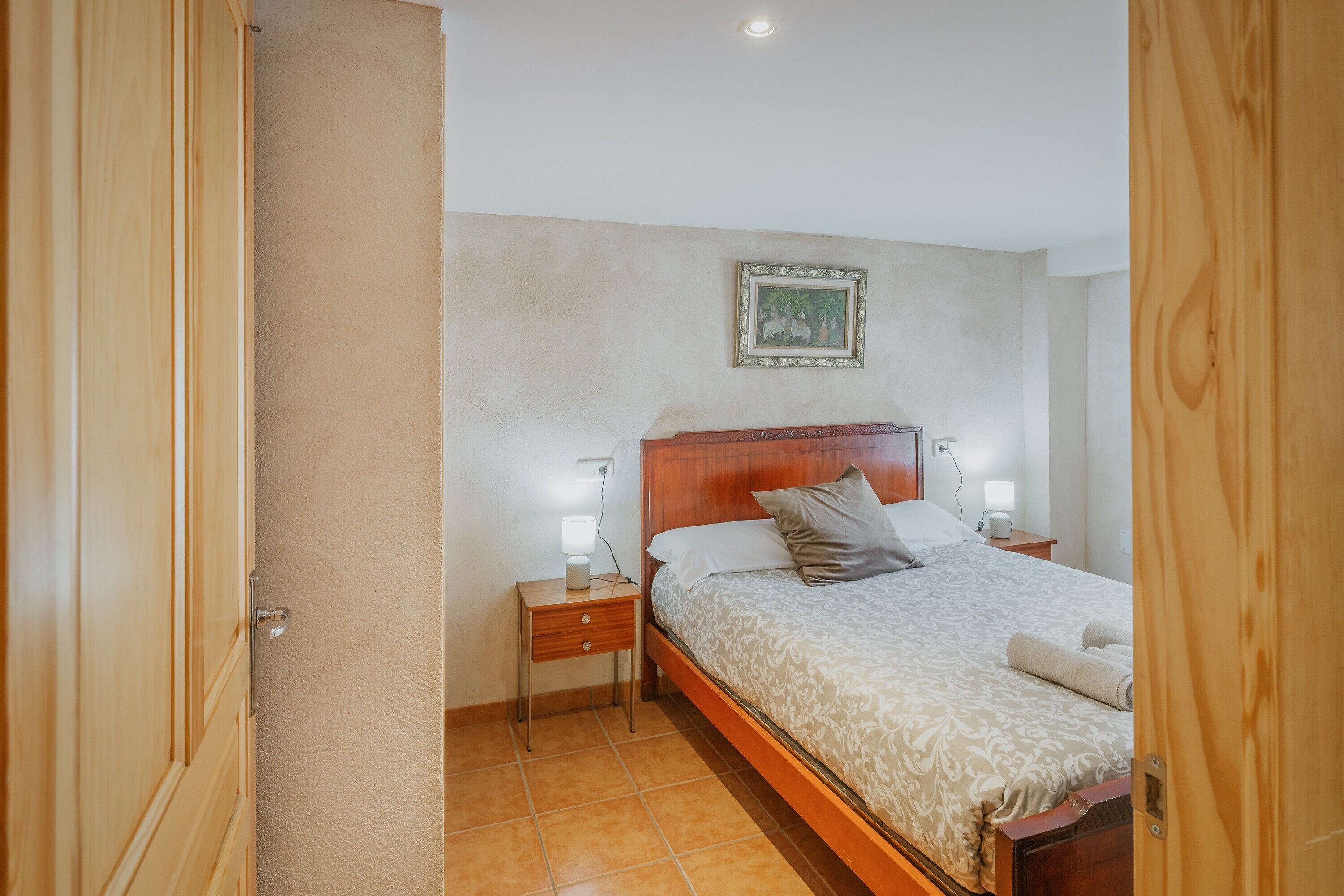Ein Schlafzimmer mit einem hölzernen Bett, Nachttischlampen, einem Nachttisch, einem gerahmten Bild an der Wand und einer teilweise geöffneten Tür.