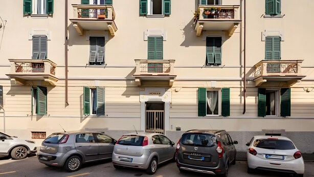 Fila di auto parcheggiate davanti a un edificio con balconi e persiane verdi.