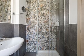 A bathroom with a white sink, a shower with a glass door, and a wall with a floral tile pattern. - Room - Rosmarino with Balcony, Wi-Fi and Air Conditioning (La Spezia)