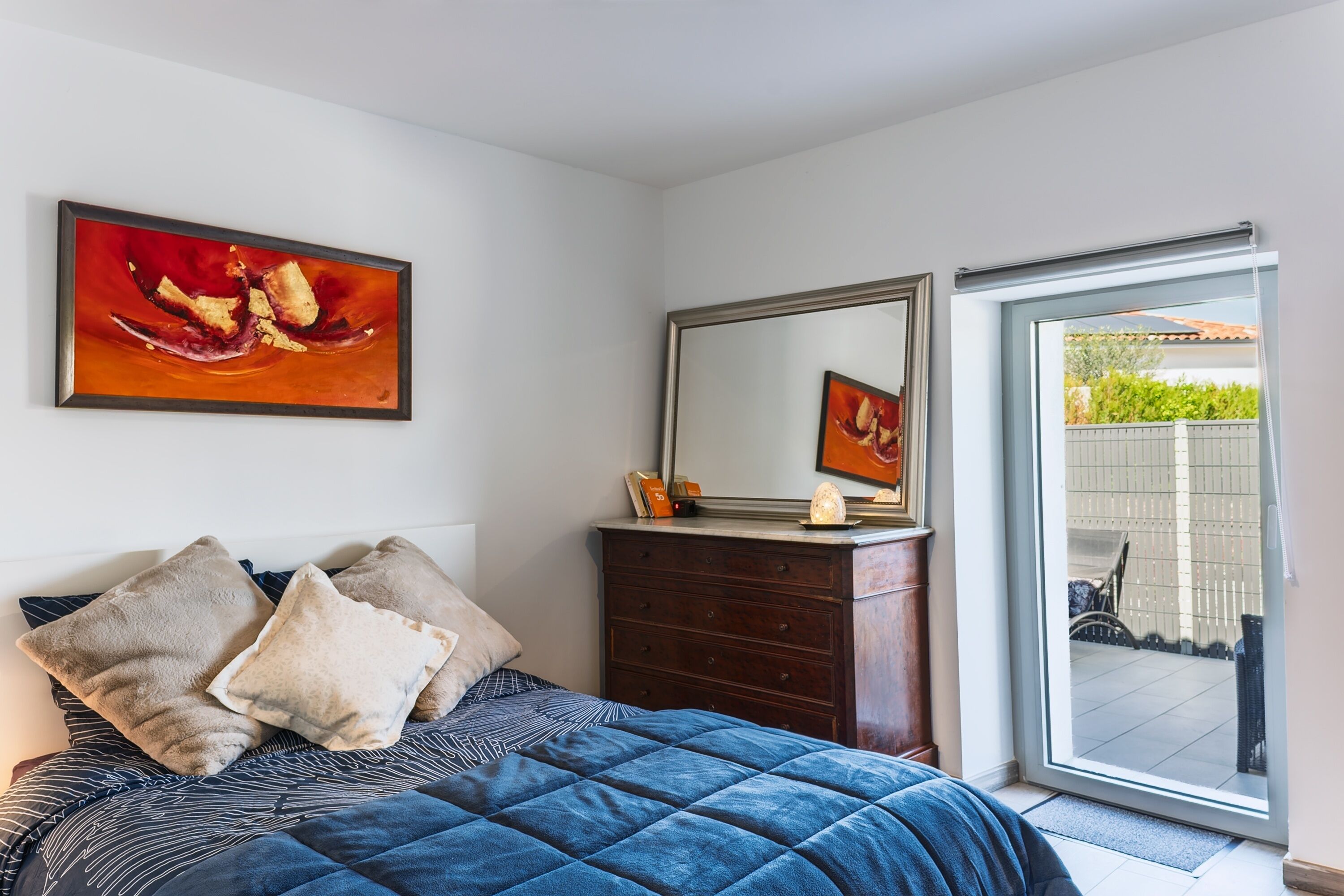 Une chambre à coucher comprenant un lit, des oreillers, une commode, un miroir et une porte-fenêtre coulissante donnant sur l’extérieur.